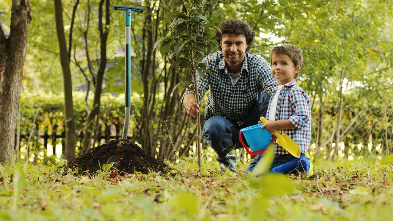 Portrait of a little boy and his dad planting a tree. They look into the camera and smile. Then the boy plays and dad - touches his son's hair. Blurred background
