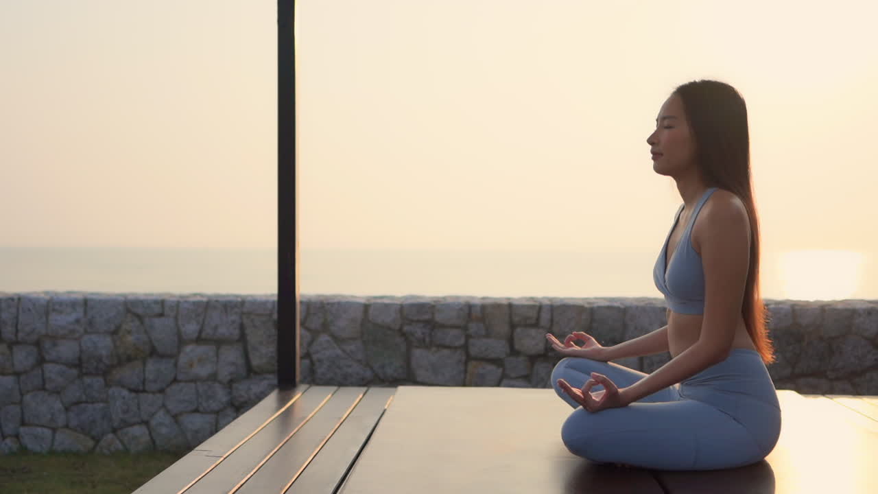 Young Asian woman in leggings doing meditation next to the sea beach ocean at sunrise, sitting in a lotus yoga pose, side view