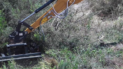 Medium-shot-on-a-windy-day-in-slow-motion-of-a-yellow-and-black-plant-machinery-arm-with-a-chain,-cutting-through-semi-dry-bushes-in-Spain