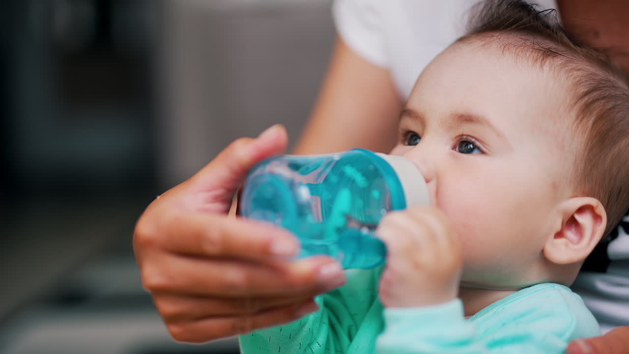 Adorable baby sits in mom's arms holding a bottle. Sweet Caucasian child drinking water. Close up.