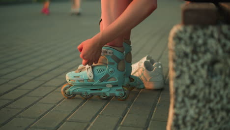 partial side view of young lady removing strap of her left leg roller skate while sitting on pavement, with two white sneakers by side and blurred legs of people walking in background