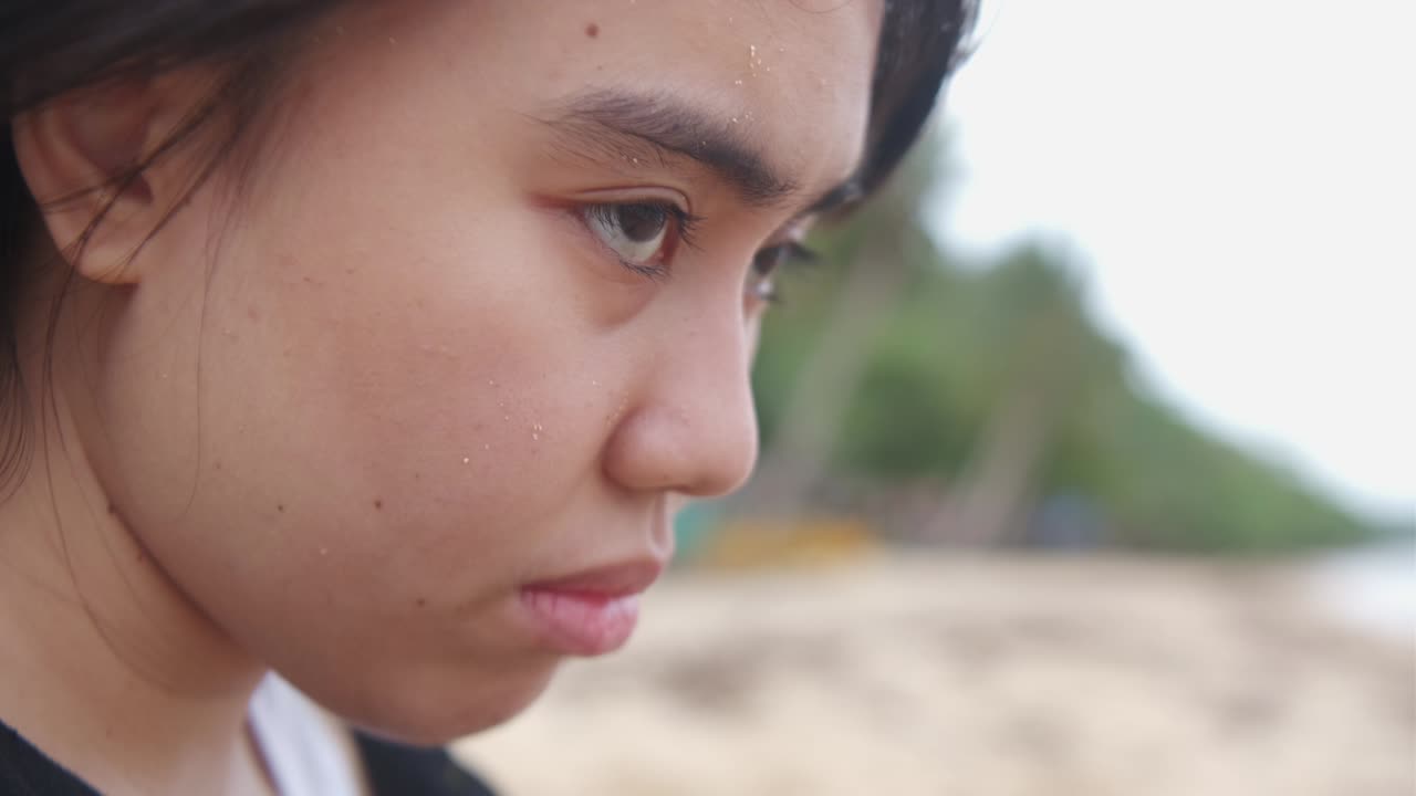 Static closeup shot of depressed young woman's face dramatically looking at horizon with wind blowing at beach setting.