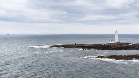 aerial camera slowly moves toward a white lighthouse on a rocky shoreline island under overcast skies, capturing tranquil seascape and gentle waves
