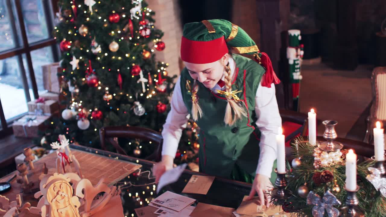 Elf girl in a green suit is looking for a letter on a table in a room with a Christmas tree and decorations