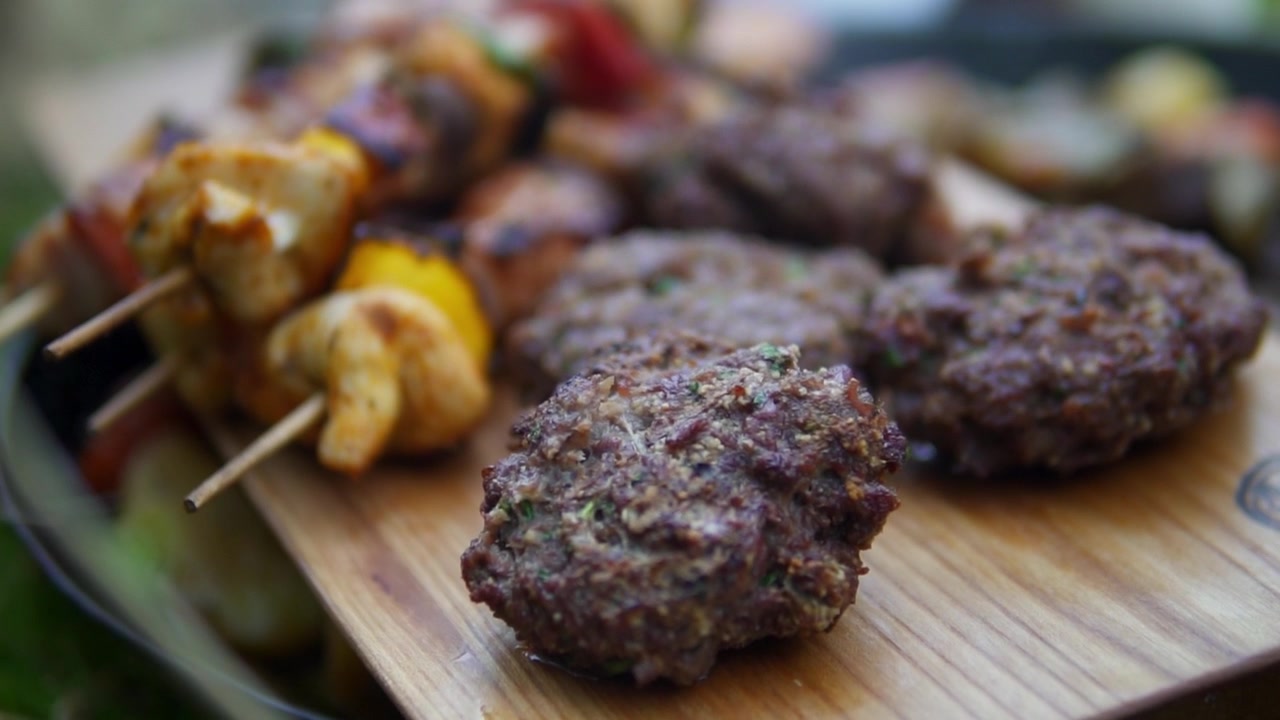 Canlı, güneşli bir yakın çekimde rustik ahşap bir tahta üzerinde duran renkli sebzeler ve burgerlerle sulu ızgara şişler. İnsan görünmüyor.