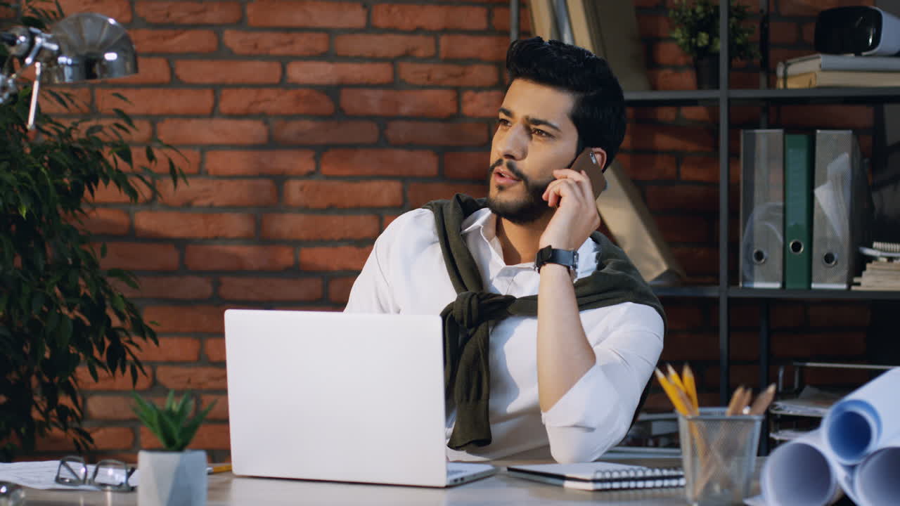 Young Arabian Businessman Sitting At The Laptop Computer Sitting At Desk And Cheerfully Talking On The Phone