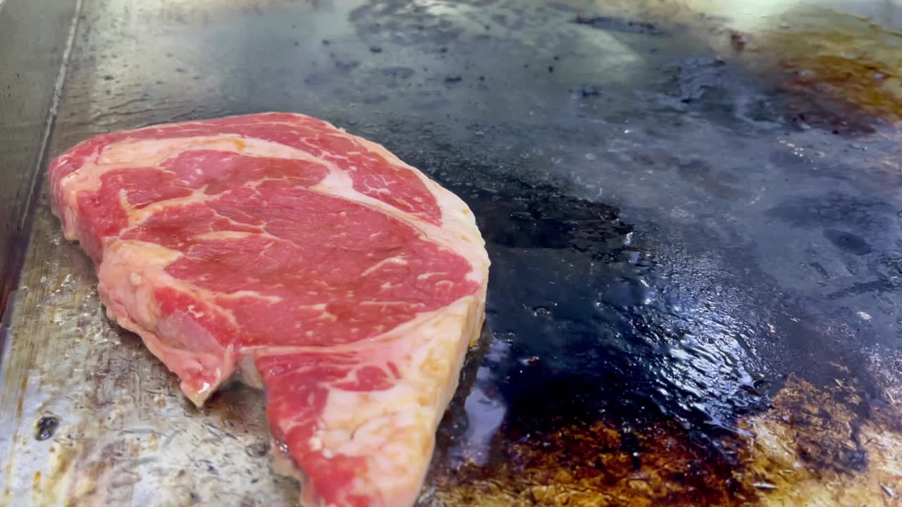 Primo piano di una bistecca di manzo cruda e fresca, posizionata su una piastra calda, che inizia a rosolare. La carne marmorizzata contrasta con la superficie di cottura scura e condita, creando un'autentica atmosfera culinaria. Idea