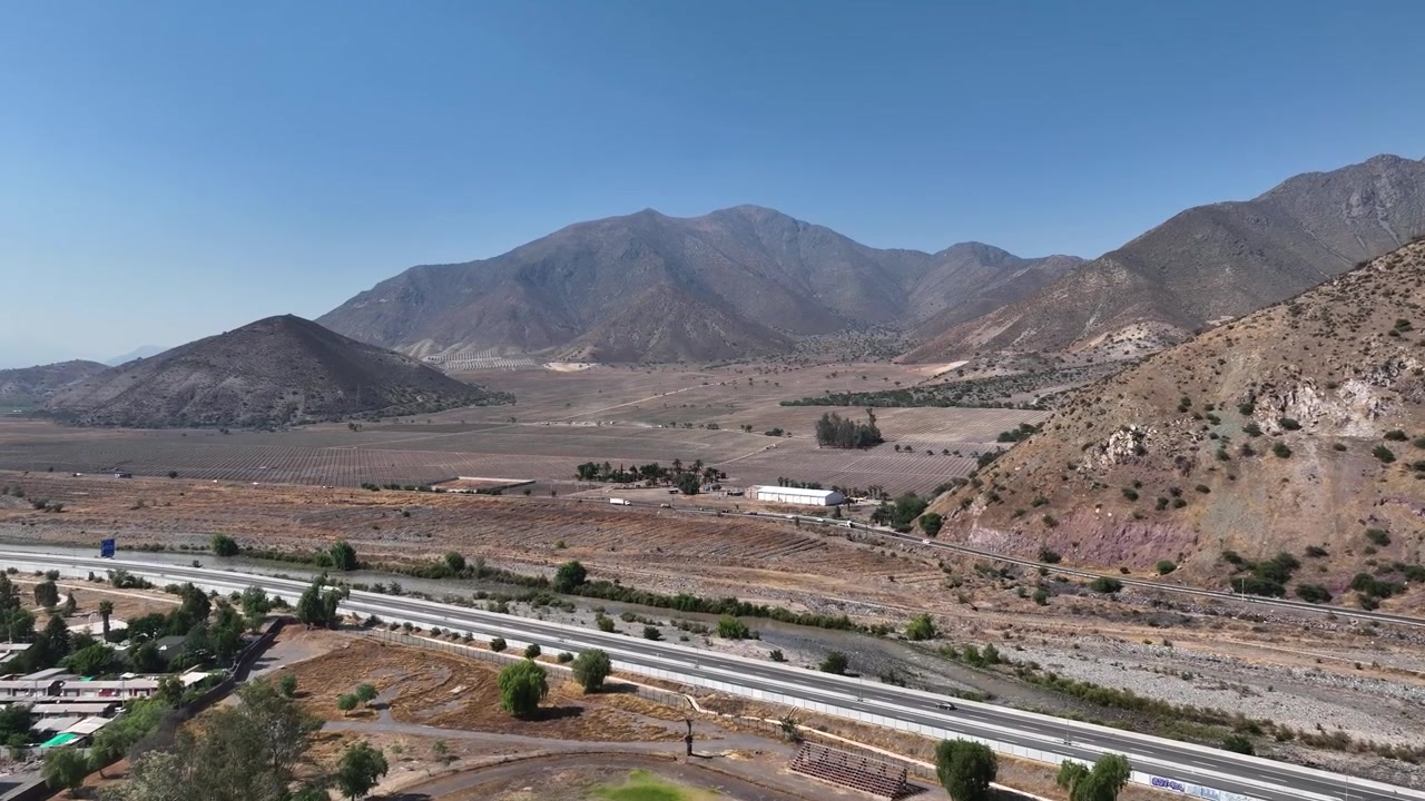 Volo in avanti sul bordo di una piccola città con condomini e impianti sportivi che rivela un vasto fondovalle arido con autostrada e drammatiche e aspre cime andine sotto un cielo azzurro e limpido.