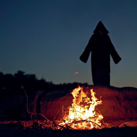 scary view of an evil in black costume at night near fire. silhouette of death with hood turning around outdoors. halloween evening.