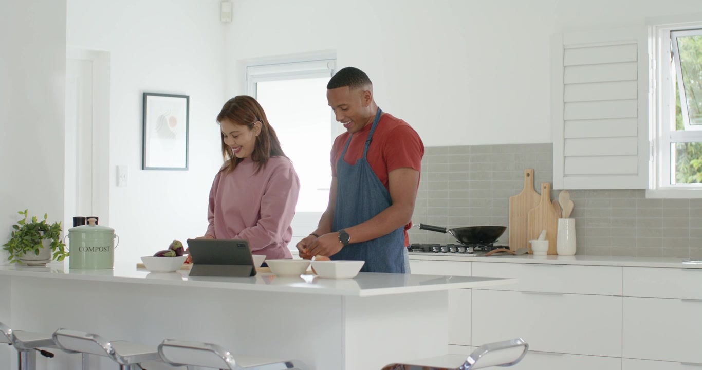 Coppia afroamericana all'isola della cucina, l'uomo in grembiule che prepara mentre la donna segue una ricetta sul tablet