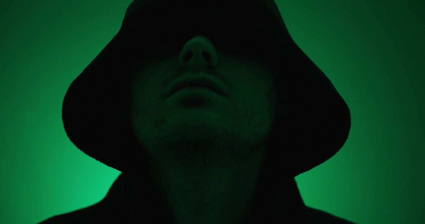 Hooded man walking slowly toward the camera under green lighting. Dramatic concept of anonymity, mystery, cybersecurity, suspense, and hidden identity.
