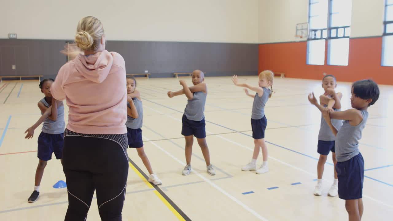 In school gym, female teacher leading group of children in stretching exercises