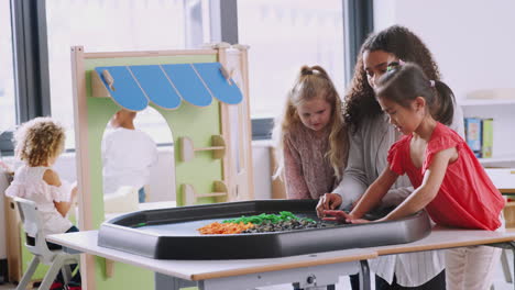 Zwei-Schulmädchen-Stehen-Mit-Einer-Lehrerin-An-Einem-Sensorischen-Spieltisch-In-Einem-Kindergartenklassenzimmer