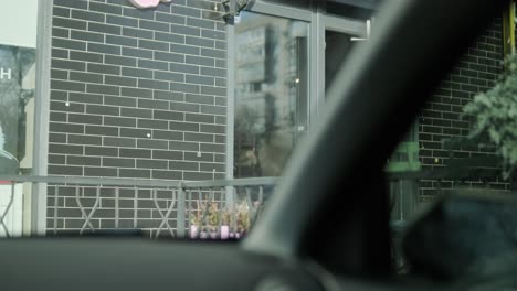 a woman dressed in a winter jacket holding a smartphone and documents stands outside a building with black brick walls. photographed from inside a car during daylight.