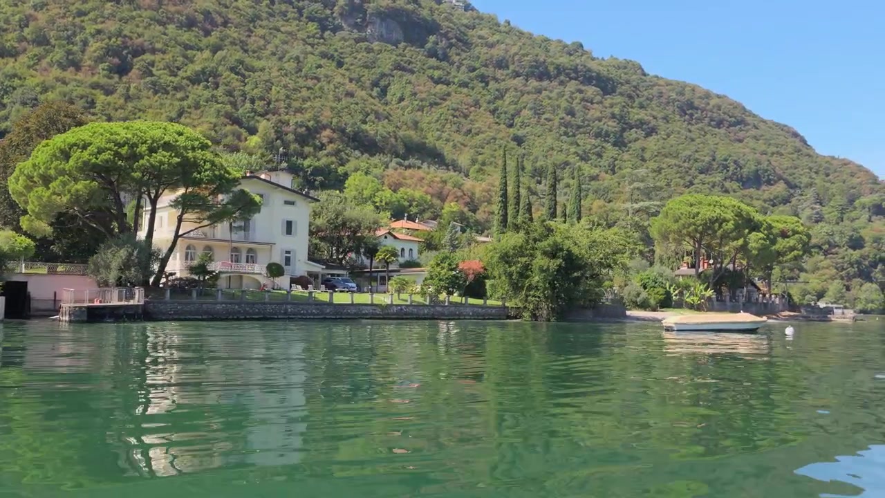 Vista serena del Lago di Lecco con rigogliose colline verdi, acqua limpida, pini ed eleganti ville sotto la luminosa luce del sole estivo.