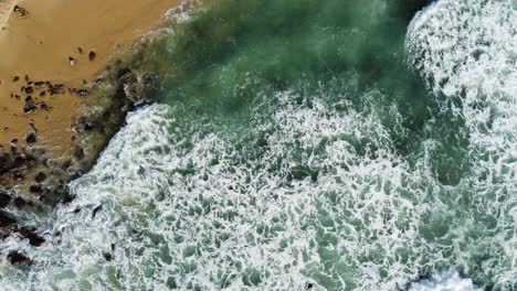 Sandy-beach-and-rocks-with-flowing-crashing-waves-above,-top-down-view