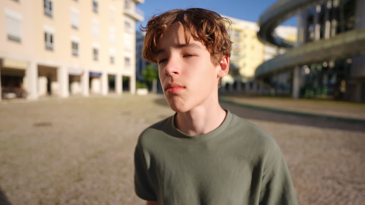 Young male teenager with curly hair stands in an urban setting, eyes closed, enjoying the sunlight, surrounded by modern architecture and a serene atmosphere