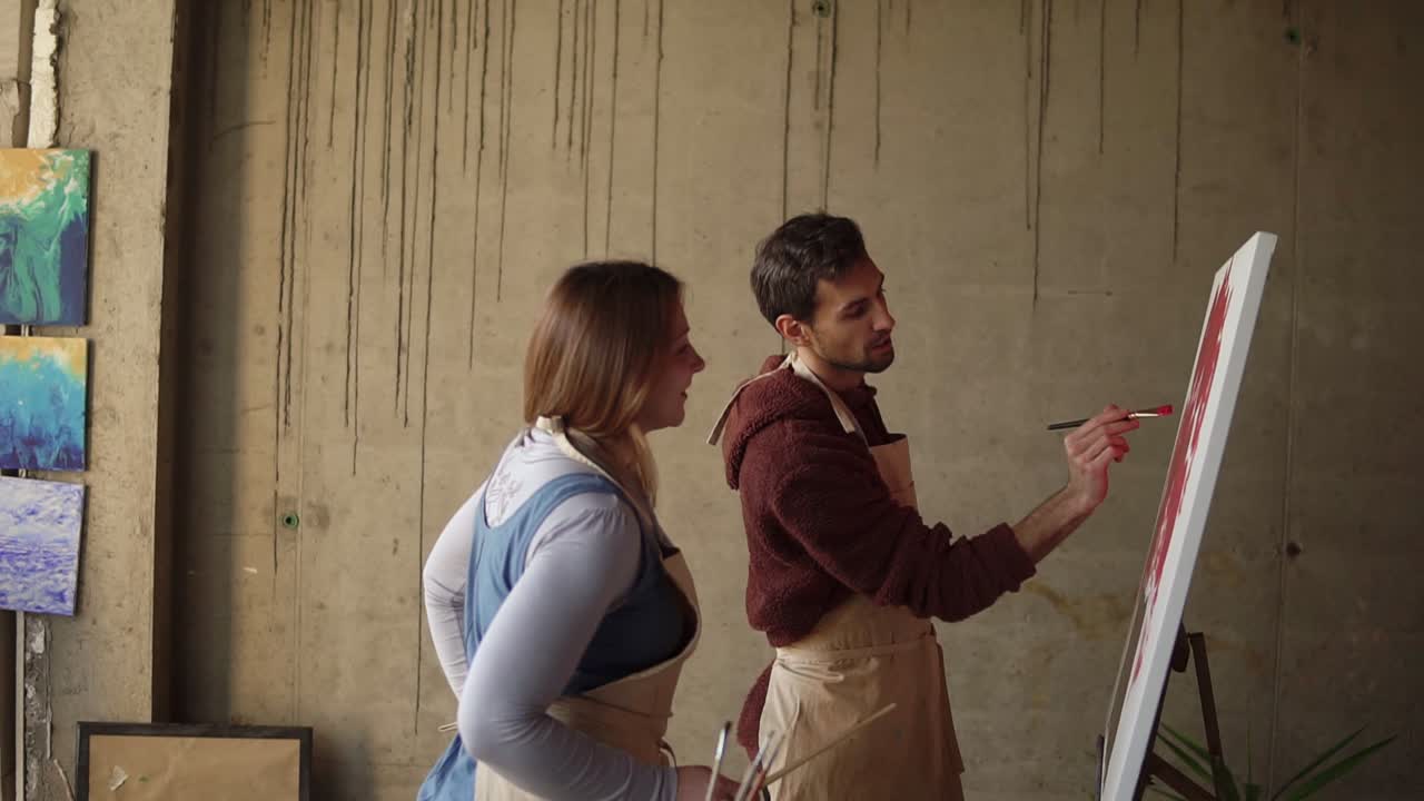 artista que enseña a un joven estudiante a pintar con pinturas al óleo. hombre de cabello oscuro con un delantal beige dibujando flores rosadas con pincel. taller en un estudio de arte
