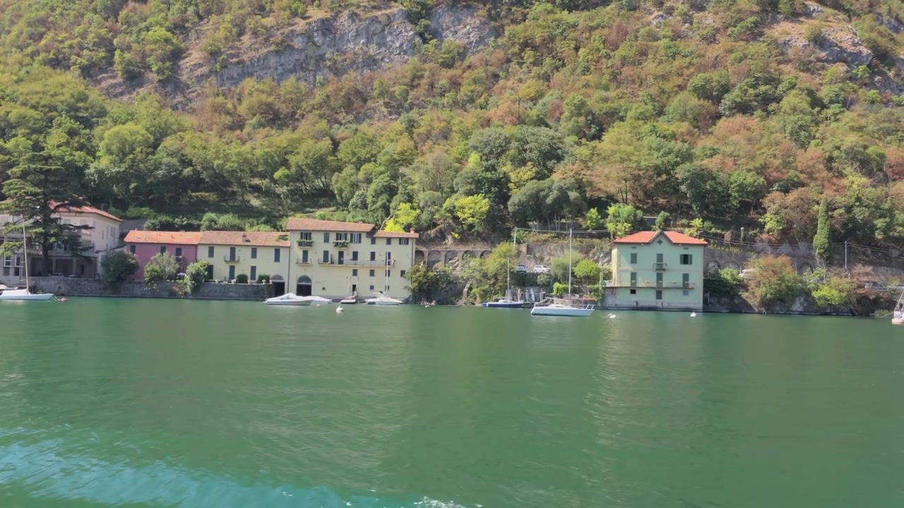 Riprese aeree che scivolano sulle tranquille acque verdi del Lago di Lecco, mostrando incantevoli ville sul lago, colline rigogliose e barche a vela ormeggiate
