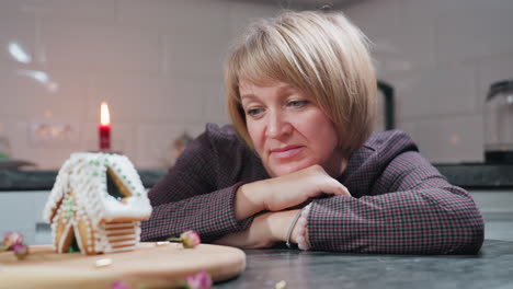 thoughtful caterer leans on table, gazing at beautifully decorated gingerbread house, she rests her head on her hands in a cozy kitchen setting with soft candlelight, reflecting on her culinary