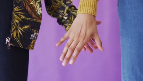 close up de una pareja diversa que se sostiene de la mano sobre un fondo púrpura