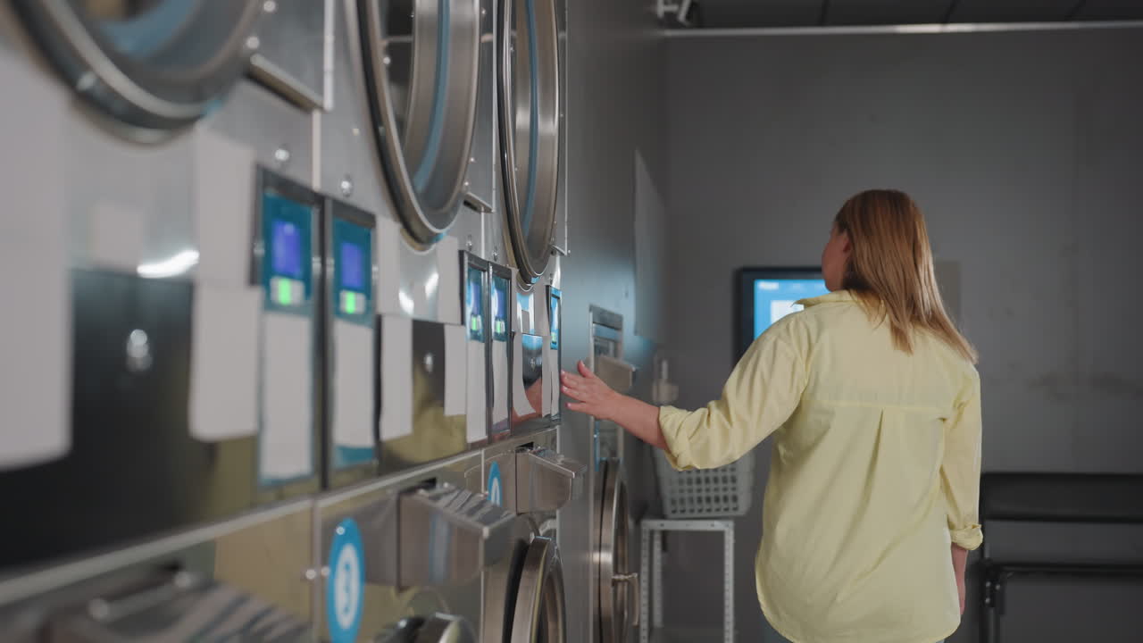 Back view employer smiling, sliding hand across industrial washers, tapping controls in modern laundromat, clothes spinning inside, basket on shelf, payment terminal glowing by corner