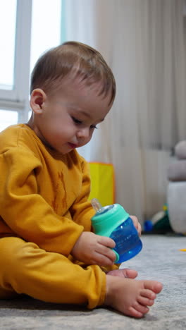 lovely barefoot kid drinks from bottle sitting on the floor. baby holds the bottle high and spills some water on himself. vertical video