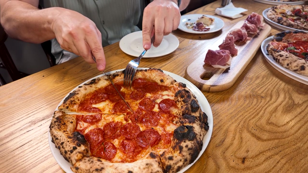 Primo piano di mani che usano coltello e forchetta per tagliare una pizza al salame piccante su un tavolo di legno.