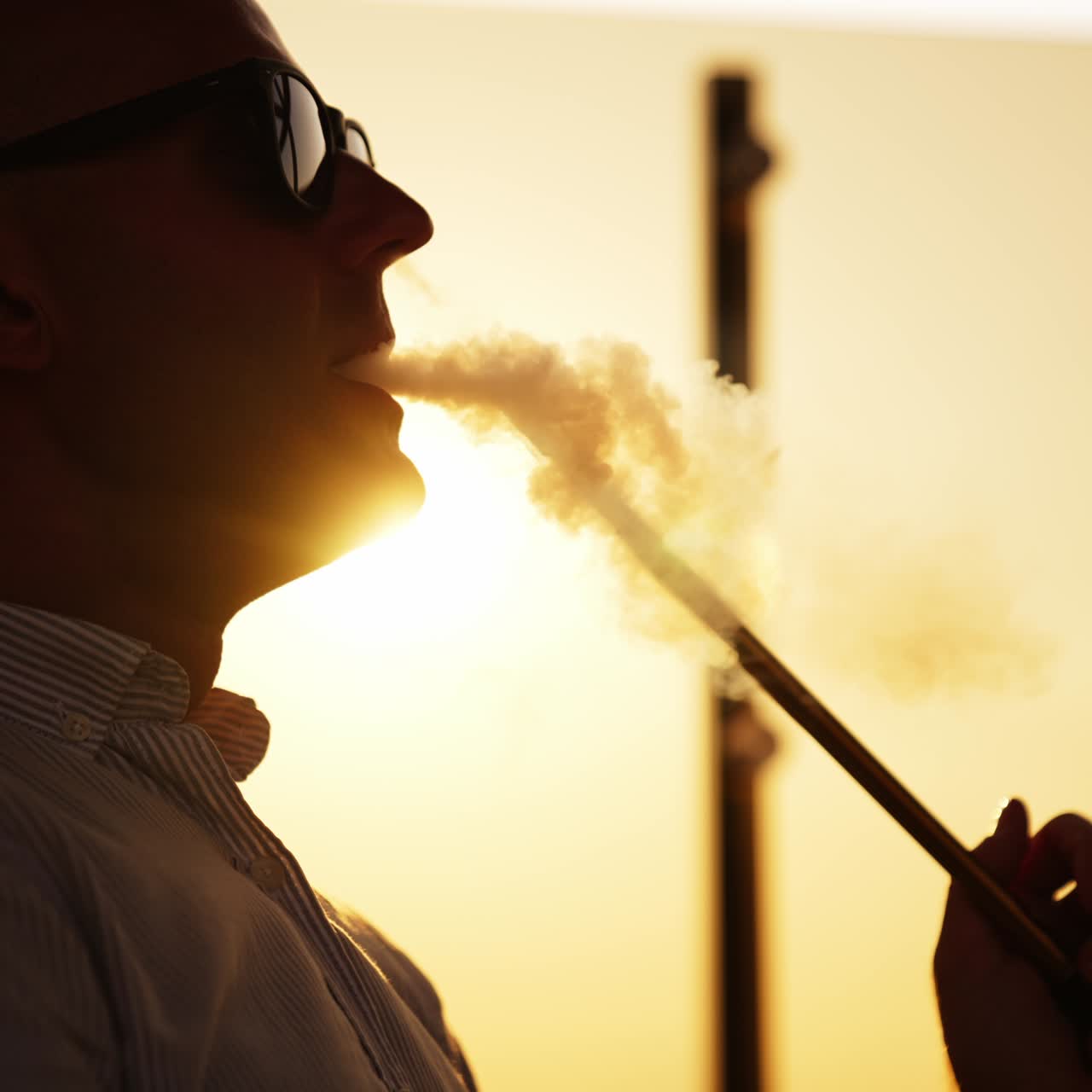 Upper part of a man in sunglasses breathing in and out thick white smoke. Young man smoking hookah at sunset close up