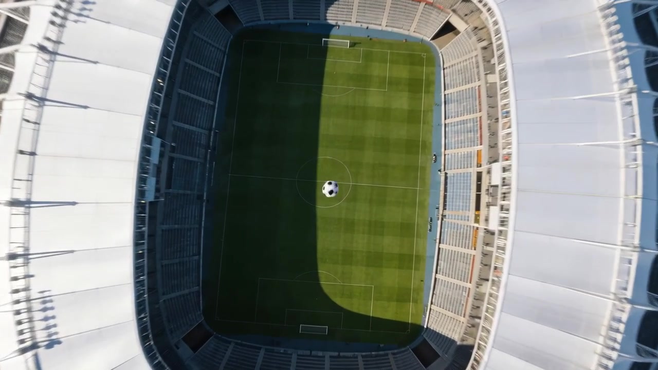 Drone in discesa che riprende un pallone bianco e nero sul cerchio di centrocampo dello stadio, mostrando le linee del campo