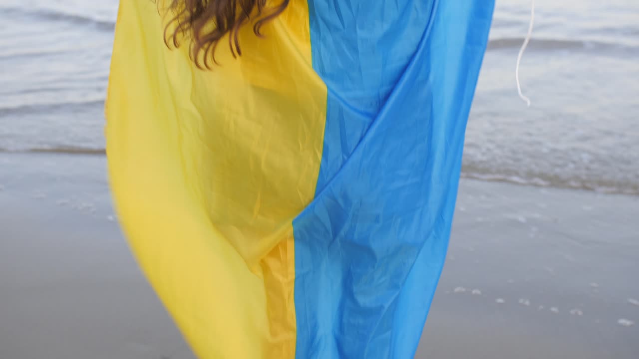 vista trasera de una chica con el pelo largo y rizado sosteniendo una bandera de ucrania sobre sus hombros descalza en la playa de arena