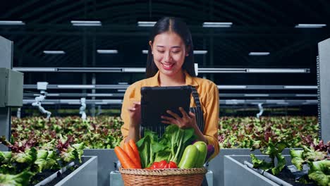 granjera asiática con una canasta de verduras sonriendo mientras usa una tableta en una granja inteligente