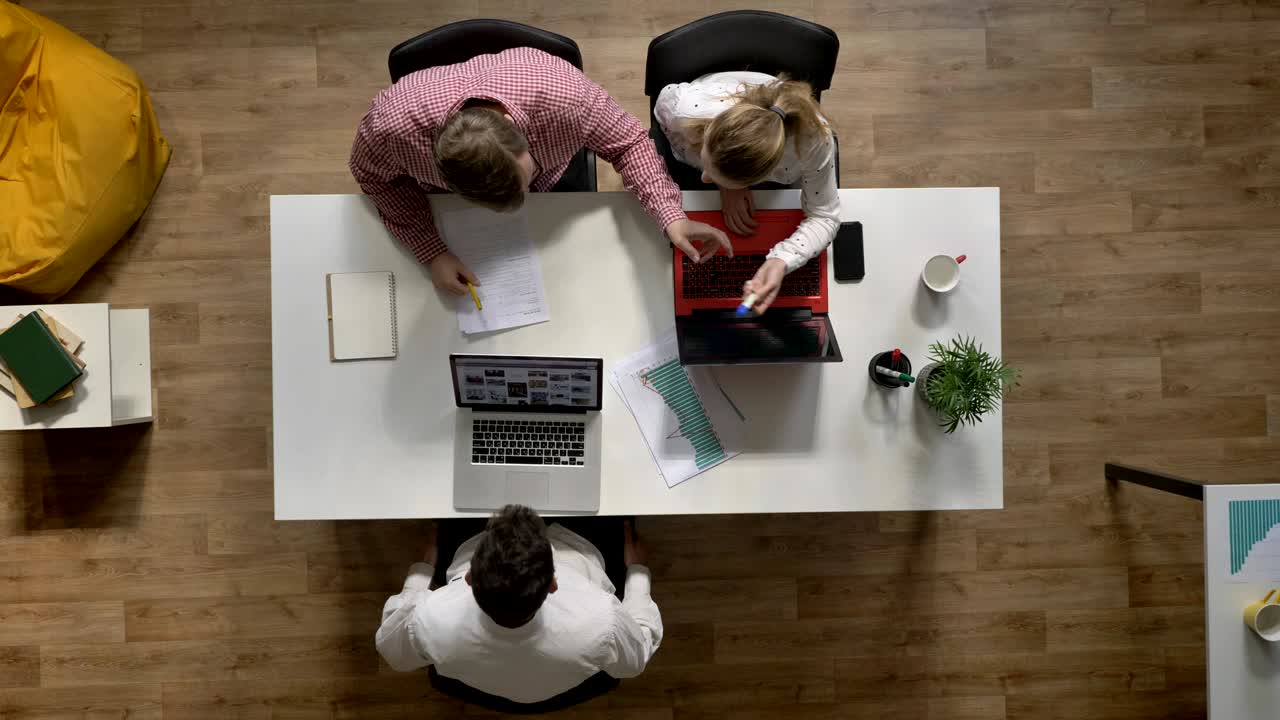 Tre unge mennesker arbejder på en bærbar computer, en anden mand skriver på et dokument på kontoret, set fra oven, siddende ved et bord og taster på en computer.