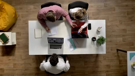 three young people working on laptop, other man writing on document in office, topshot, sitting at table and typing on computer