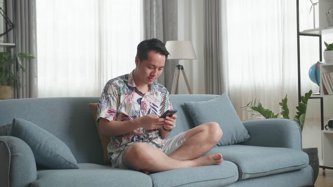 Asian Man Sitting On Couch In Living Room And Using Smartphone