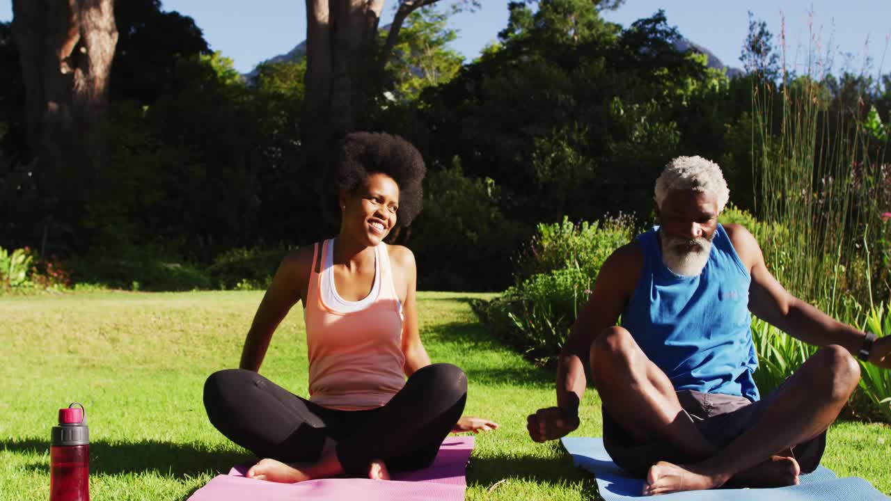 Casal idoso afro-americano praticando yoga sentados em um jardim ensolarado.