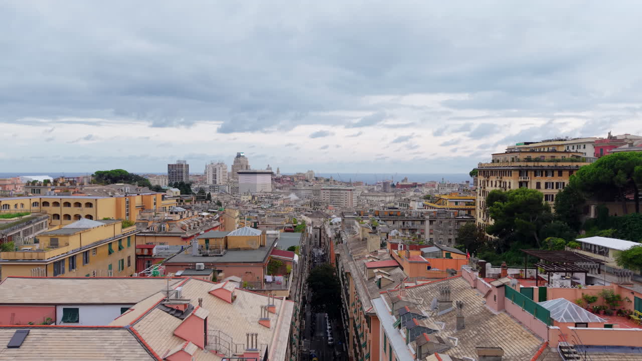 The drone flies above the rooftops of Genoa, revealing colorful old buildings, the city skyline, and the sea in the distance under a cloudy sky