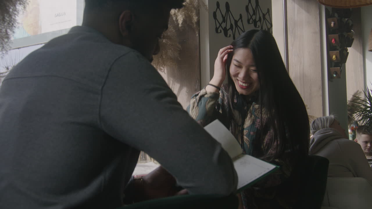 Multi Ethnic Couple Watch Menu and Talk Multi Ethnic Couple Sit at the Table Near Window Watch Menu and Talk African American Man and Asian Woman on a Date in Cafe Diverse Guests in Restaurant with Modern Design Public Eating Concept