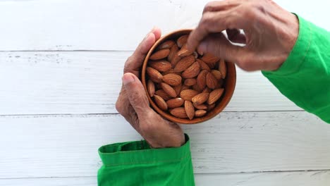 manos envejecidas recogiendo almendras de un cuenco de madera en una mesa blanca