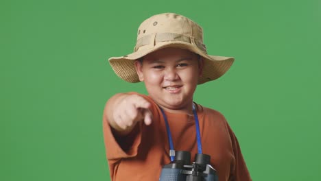 niño turista asiático con sombrero y binoculares sosteniendo una lupa, tocando su pecho y luego señalándote en el fondo de la pantalla verde. niño investigador examina algo, turismo de viajes, de cerca