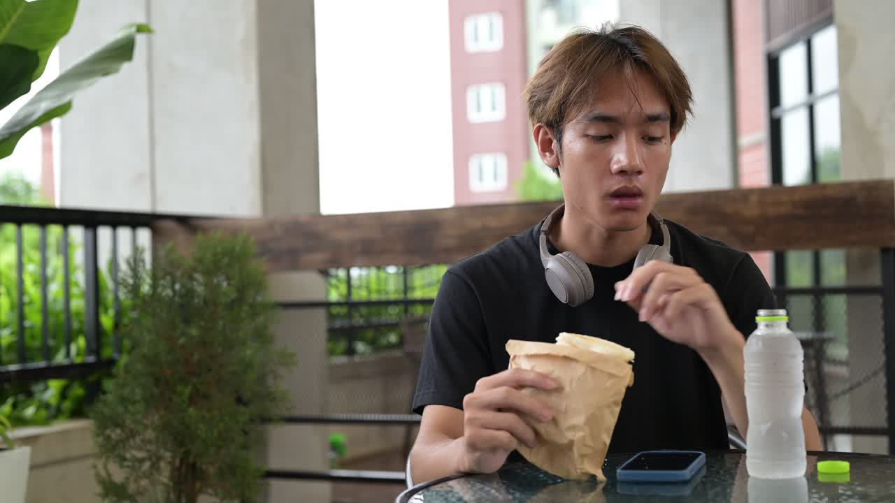 Happy young Asian Thai student man eating sandwich and drinking water outdoors for casual lunch while sitting alone