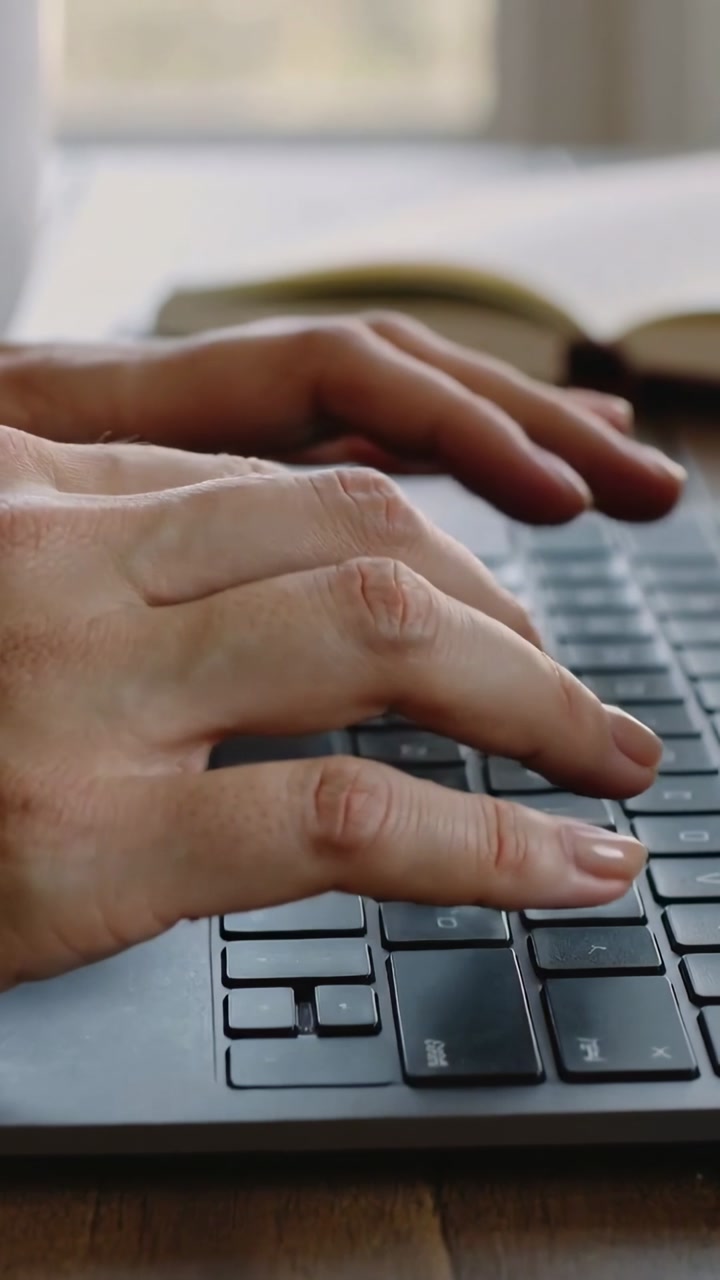 Vertical video: Resting hands pressing keys on laptop at desk, working on text, book blurred