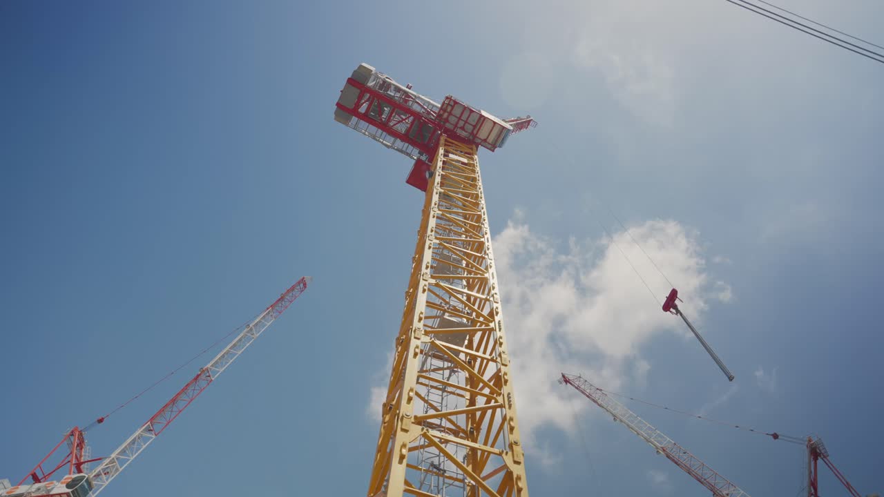 Yellow tower crane from below, Construction site.reaching into a bright blue sky with thin clouds. More cranes edge the frame. development, infrastructure, real estate. Upward. Ambitious. Expanding