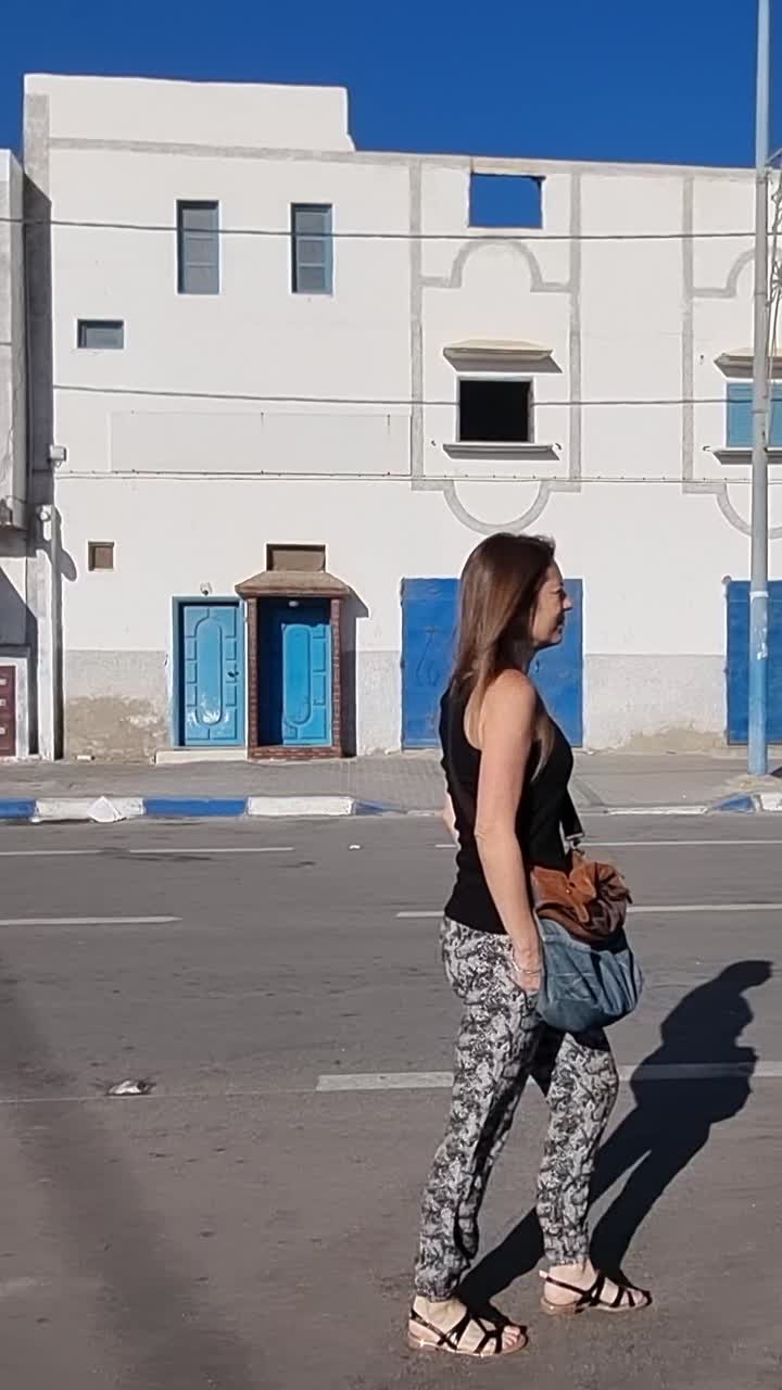 Donna che cammina per una strada a Tarfaya, Marocco. Giornata di sole con un edificio tradizionale bianco e blu sullo sfondo.