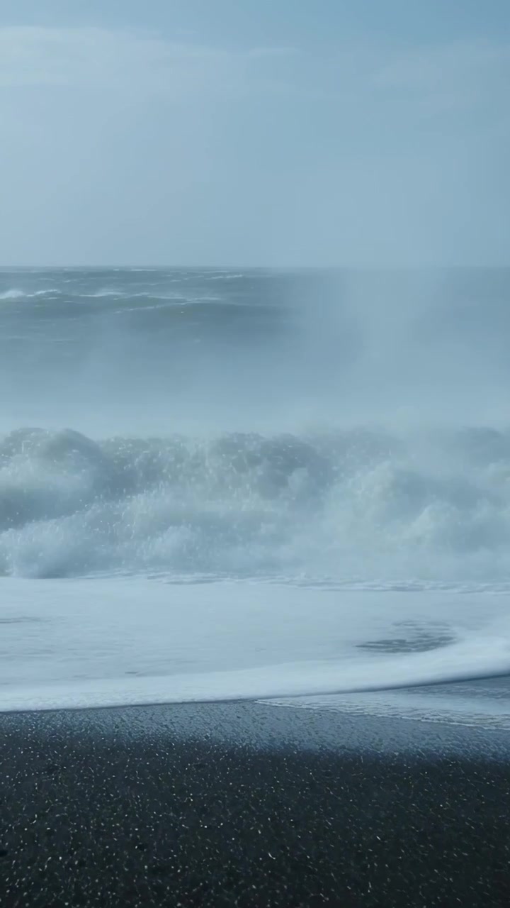 Video verticale: Onde oceaniche che si infrangono su una spiaggia di sabbia nera mentre arrivano le mareggiate, generando schiuma.