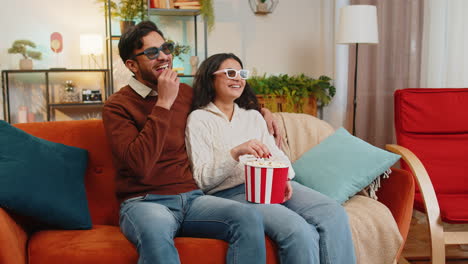 happy indian family couple in 3d glasses eating popcorn and watching comedy movie sitting on sofa