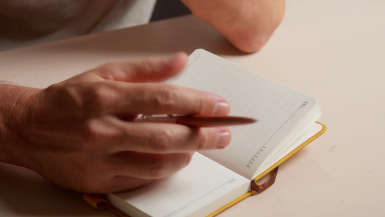 Man sits with notebook and pen at an indoor table. He is struggling with what to write and is distracted and procrastinating. Clip 1