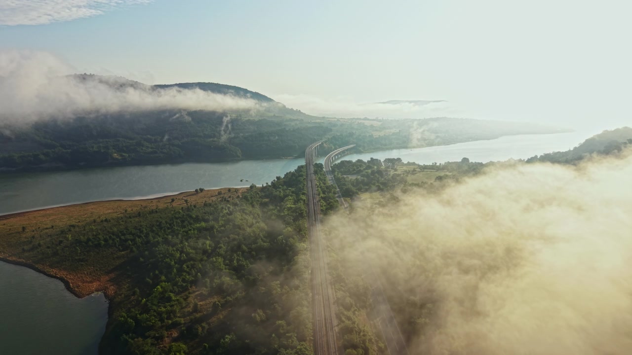 Aerial fog drifting over wooded peninsula into sheltered bay with soft morning sunlight revealing treetops, calm water, layered shoreline, and distant coastal haze