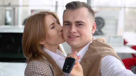 happy couple taking a selfie at a car dealership with a new car in the background. man and woman showing keys of his new car in dealership