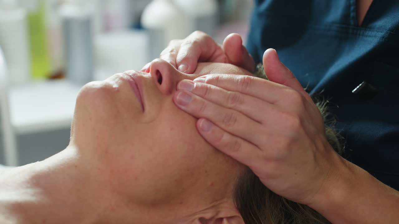 High angle close up of skin care expert placing hands on reclined woman on massage bed and gently massaging eyelid and surrounding eye area with smooth strokes under bright spa lighting